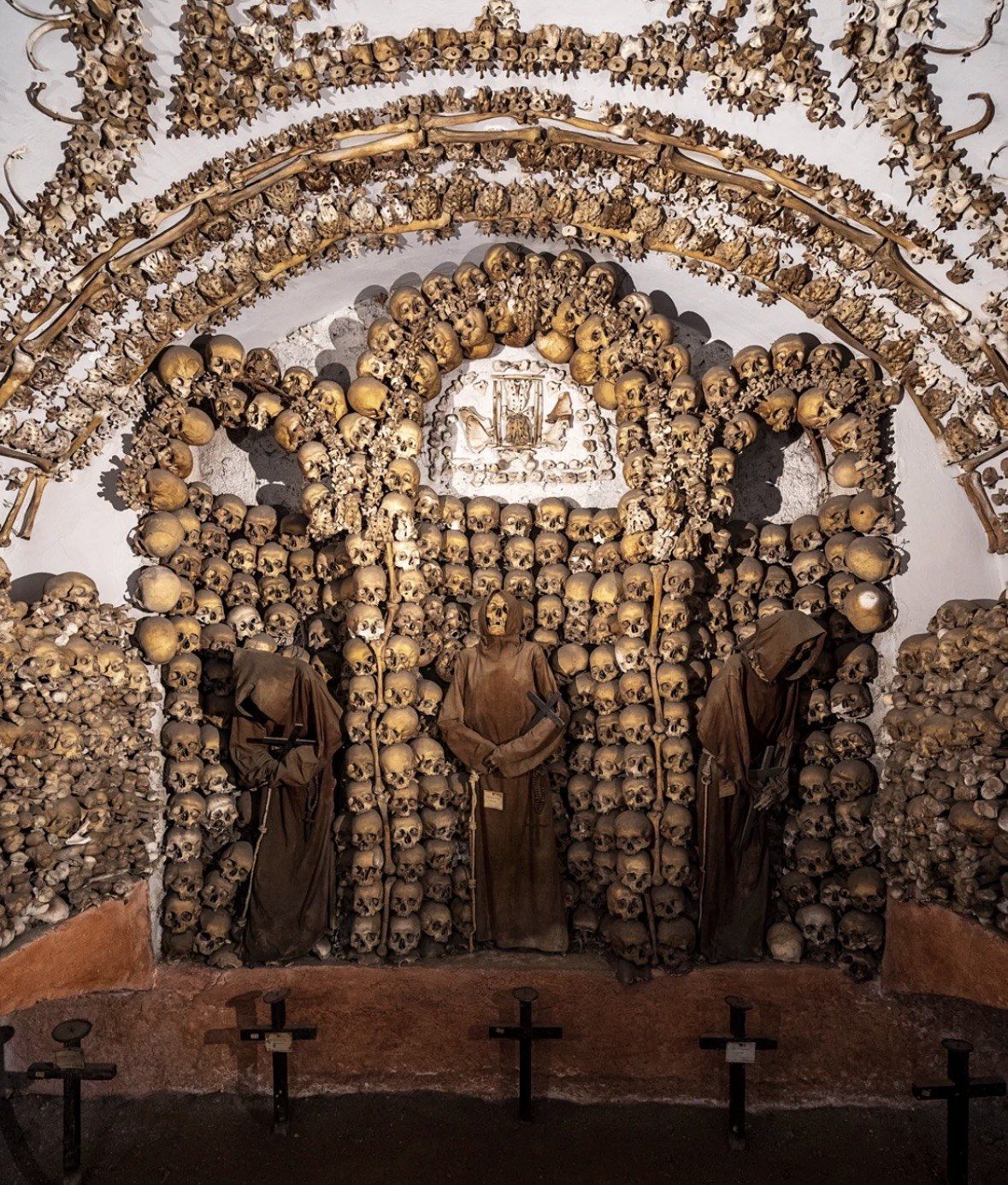 Three robed friars standing under an arch built from skulls and bones